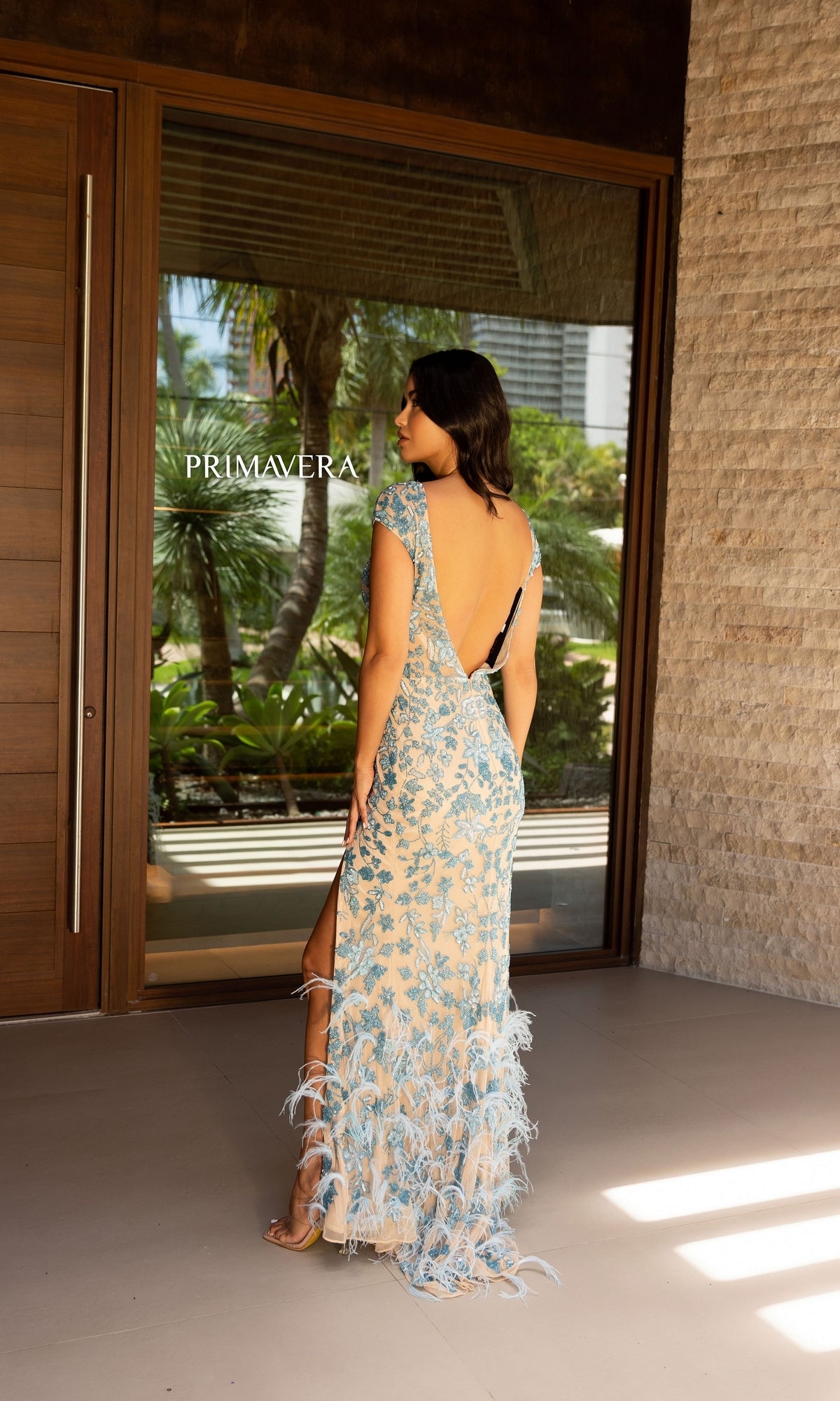A woman stands indoors, facing away, wearing the Feathered Cap-Sleeve Long Beaded Prom Dress 12107 with a low back, as palm trees are visible through the window behind her.